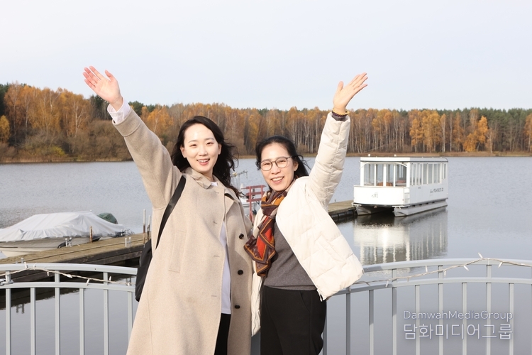 자슬라브스코예 호수(‘민스크 바다’) 선착장에서 김선아 『Diplomacy Journal』 특파원 기자가 어머니와 함께 환하게 손을 들어 인사하고 있다. 가을빛으로 물든 단풍과 잔잔한 호수 위 유람선이 배경을 이루며, 모녀의 밝은 미소가 벨라루스의 평화로운 풍경 속에 따뜻하게 녹아든다.